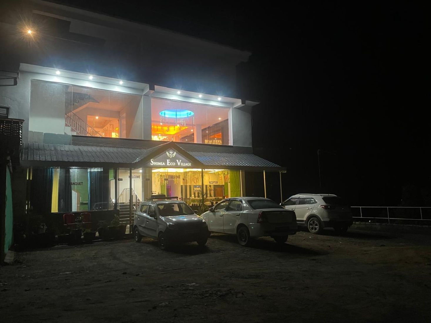 A two-story building with a sign reading 'Shima Eco Village.' There are cars parked in front, and the entrance is visible.