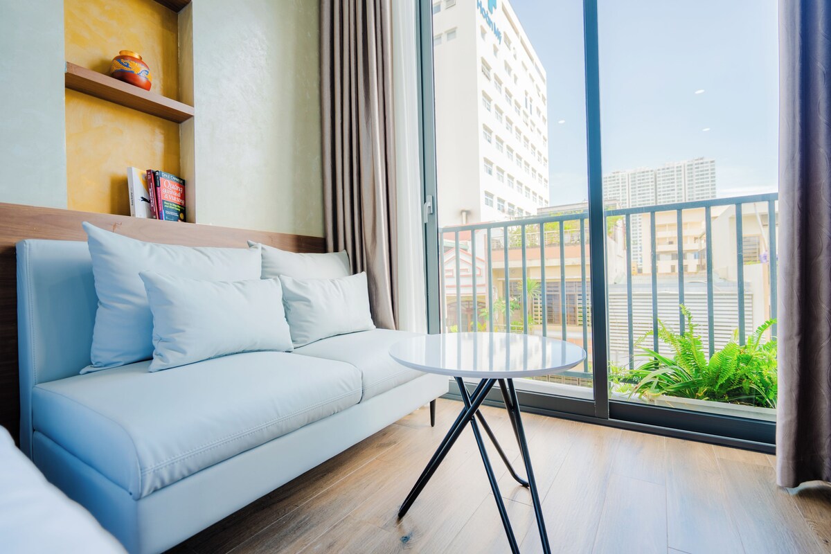 Una habitación de hotel moderna con un sofá azul claro, una mesita redonda y vistas de la ciudad a través de amplias ventanas.