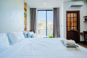 Una habitación de hotel con una cama, una mesita con libros, una ventana con cortinas y un equipo de aire acondicionado.