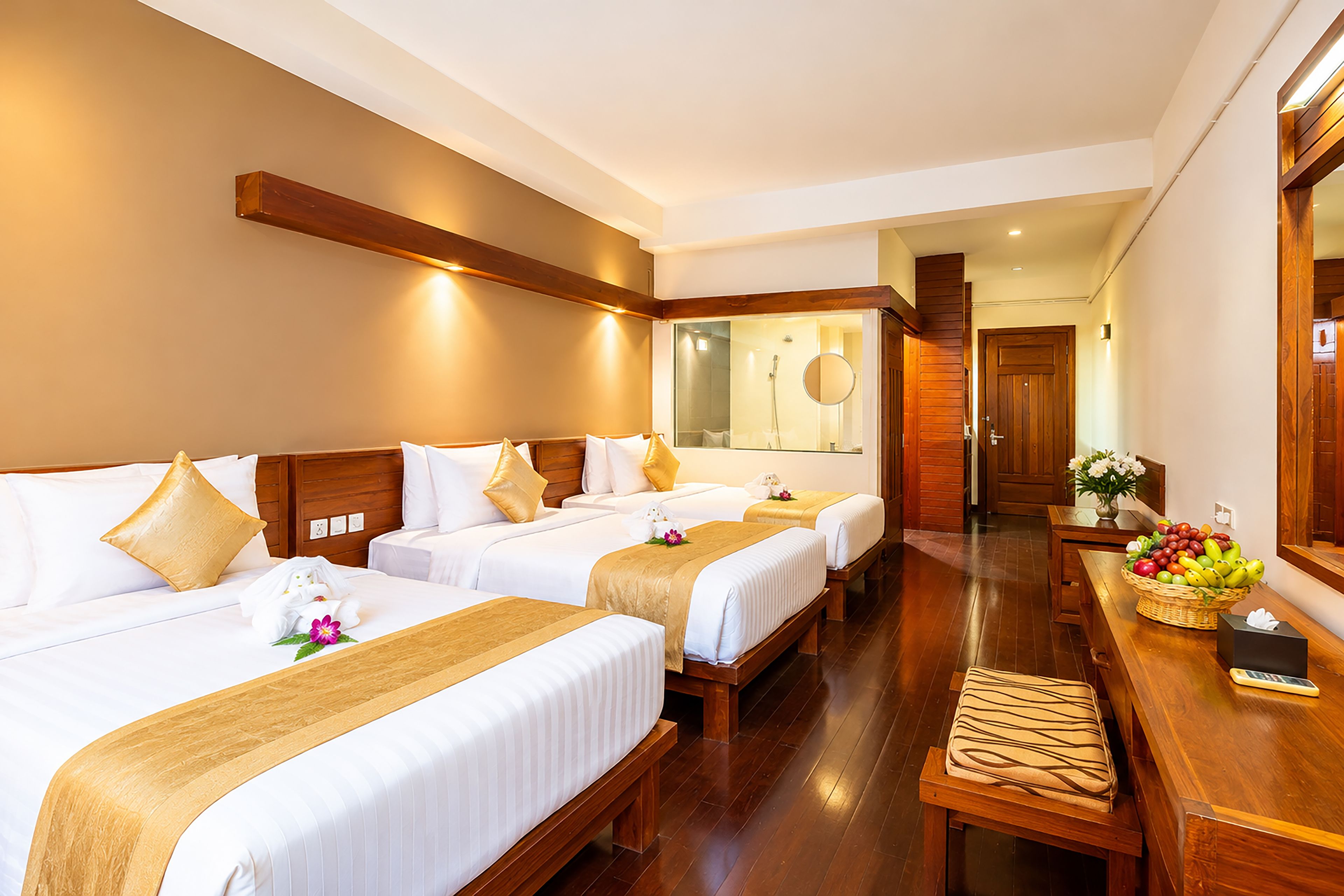 A hotel room with two beds, a wooden bench, a desk with a basket of fruit, and a mirror.