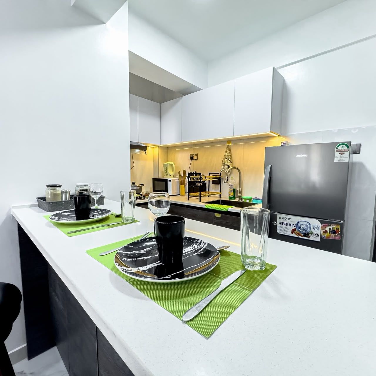 Een moderne keuken met witte kasten, een roestvrijstalen koelkast en een eethoek met een witte tafel gedekt voor twee personen.