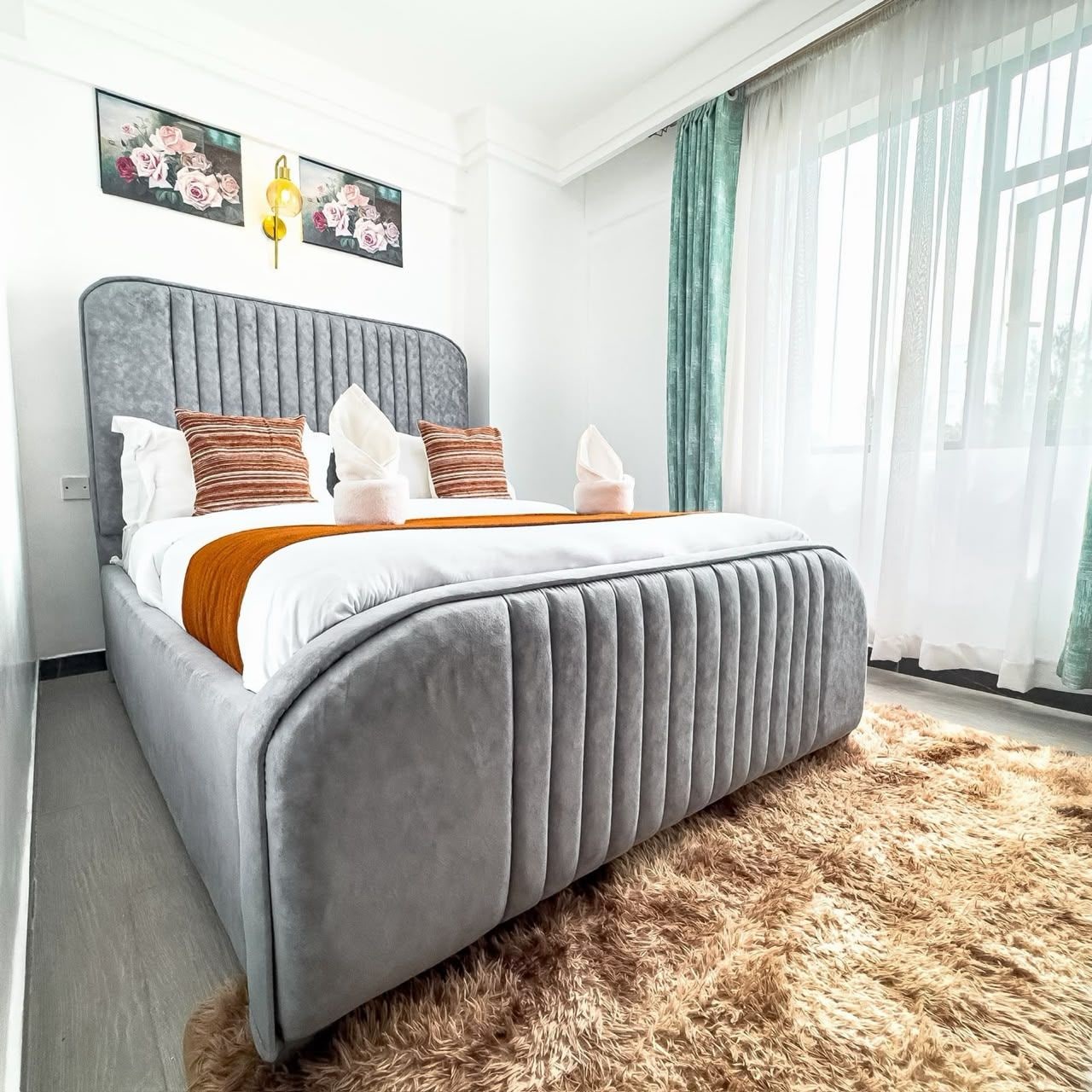 Een moderne slaapkamer met een gestoffeerd hoofdeinde, een pluizig vloerkleed en een groot raam met dunne gordijnen.