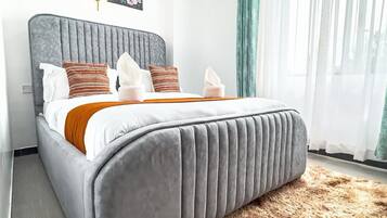 Een moderne slaapkamer met een gestoffeerd hoofdeinde, een pluizig vloerkleed en een groot raam met dunne gordijnen.