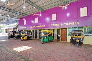 A building with a purple facade advertising 'TOURS & TRAVELS' and 'ROOMS AVAILABLE'. There are auto-rickshaws parked in front. - Hotel O PULARI BACKWATER & ROOMS (Neyyattinkara)