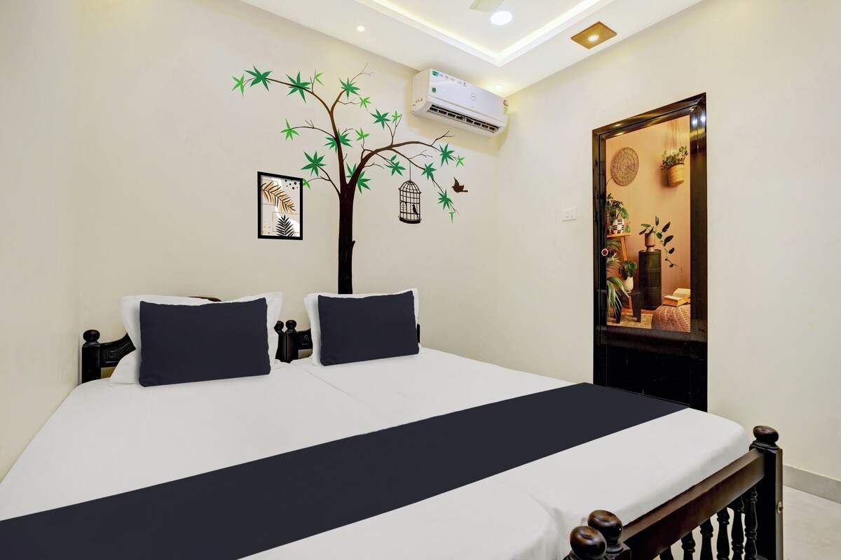Quarto de hotel com cama, ar-condicionado fixado na parede, porta que dá para um ambiente com plantas e decoração de árvore com pássaros.