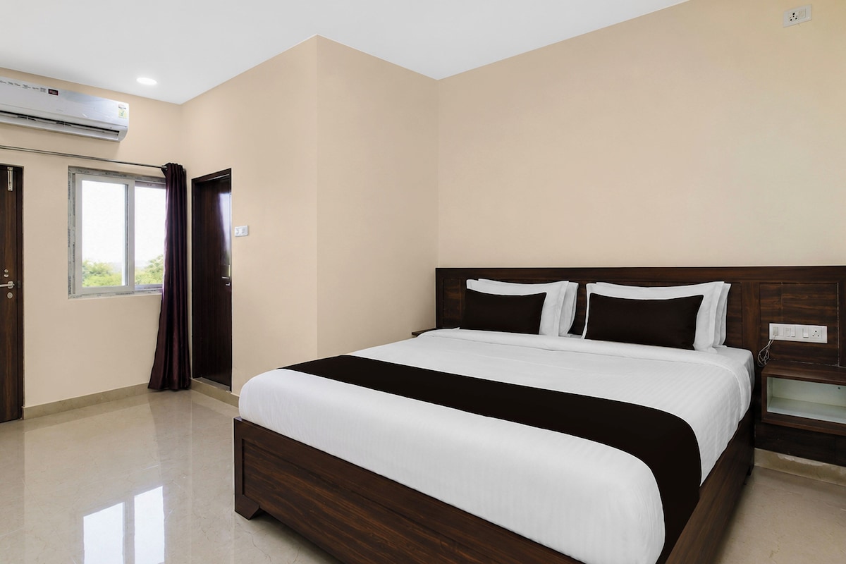 A hotel room with a large bed, a bedside table, a window with curtains, and an air conditioning unit.