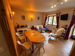 Een gezellige woonkamer met een houten eettafel, witte stoelen, een grijze bank en een aan de muur bevestigde televisie.