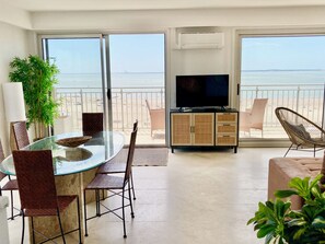 Ein modernes Wohnzimmer mit einem Glastisch, Korbstühlen, einem Flachbildfernseher und Blick auf den Strand.