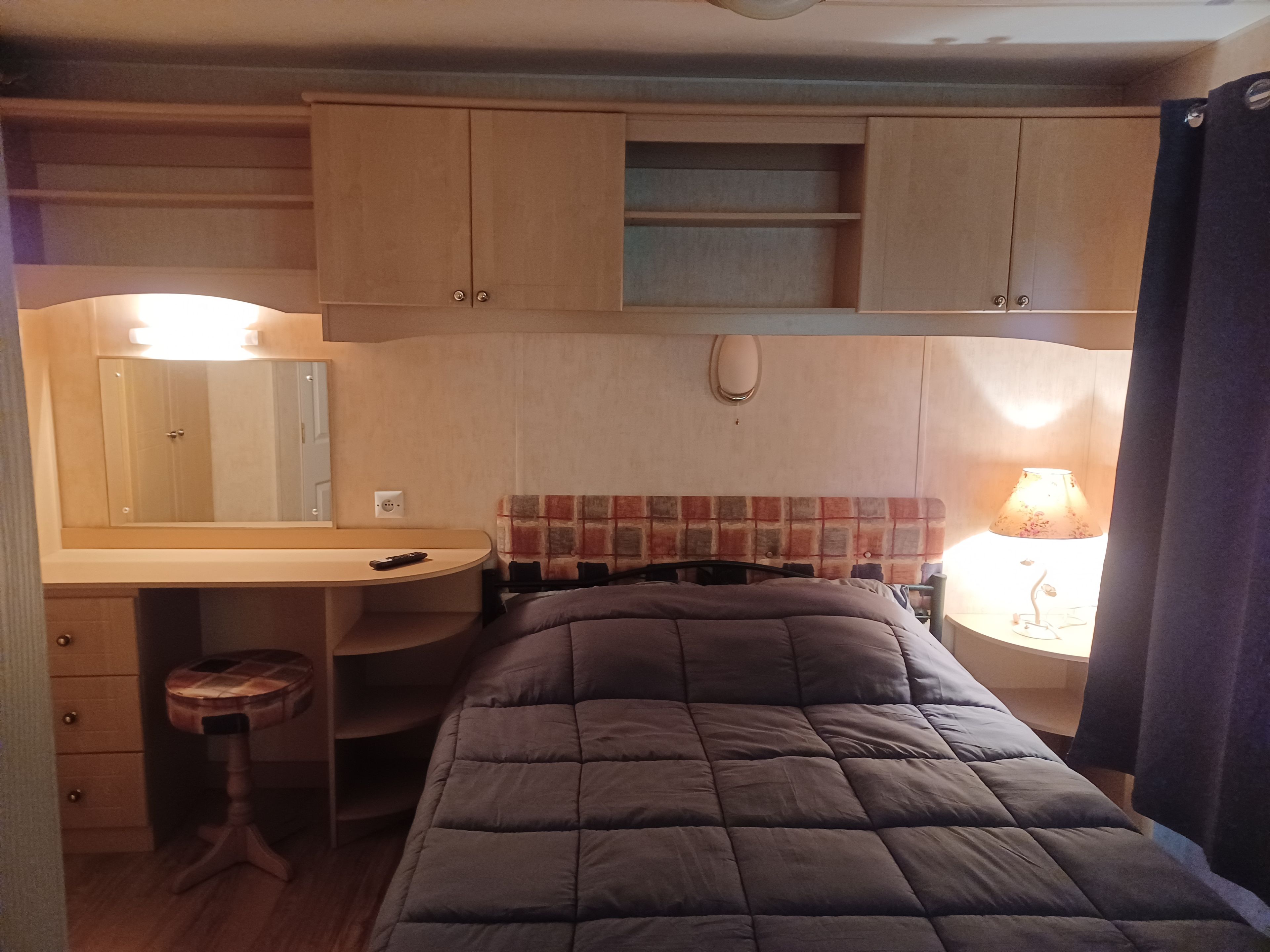 A compact bedroom with a bed, a small desk, a chair, a mirror, and a bedside lamp.