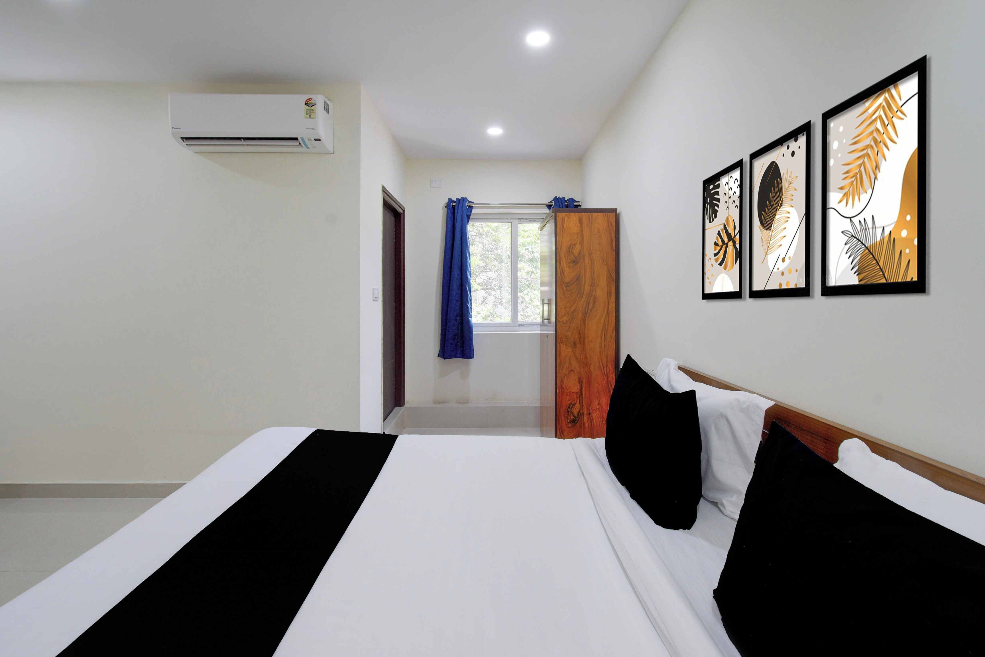 A hotel room with a bed, a window with curtains, an air conditioning unit, and framed artwork on the wall.