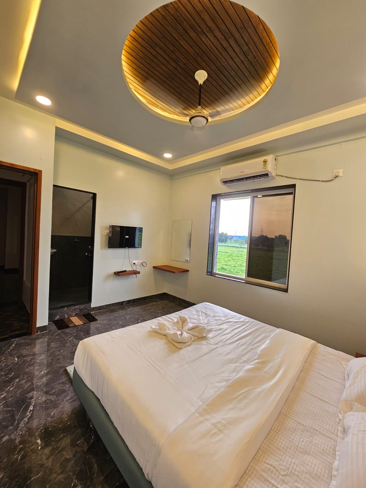 Ein Hotelzimmer mit Bett, Deckenventilator, Wandfernseher und einem Fenster mit Blick auf Grünflächen.