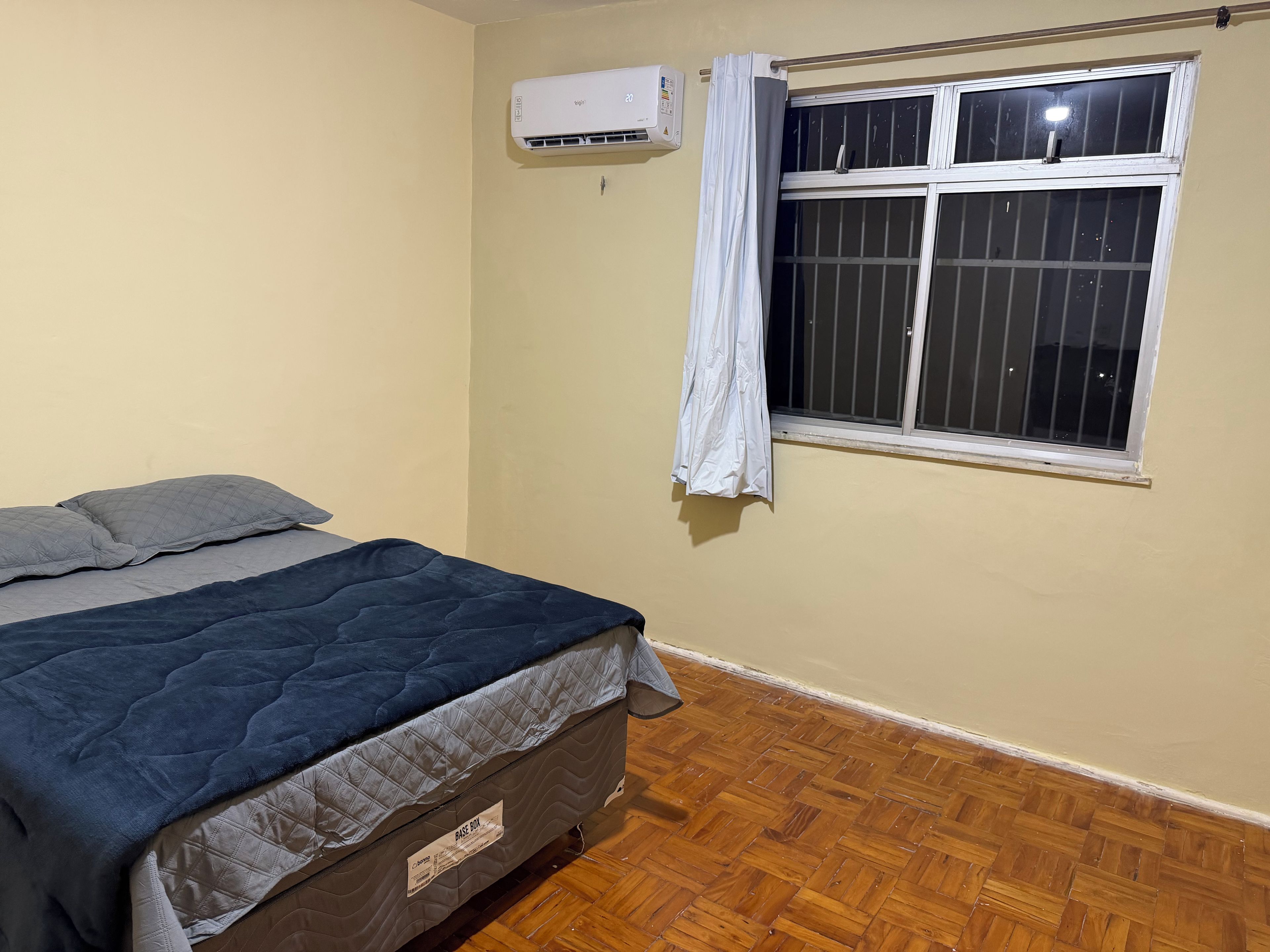 Une chambre à coucher avec un lit, une fenêtre avec des barreaux, un appareil de climatisation et un plancher en bois.