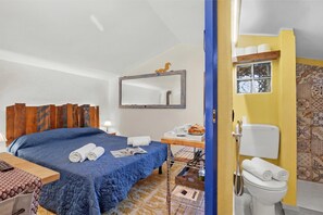 Habitació d'hotel amb un llit, una taula de nit, un mirall, un petit escriptori amb una cadira, i un bany amb vàter i dutxa.