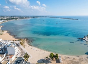 Une vue aérienne d’une zone côtière avec une plage, des eaux d’un bleu limpide et des bâtiments le long du rivage.