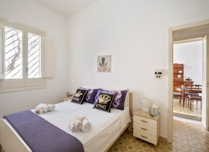 Une chambre à coucher comprenant un lit, des oreillers, une table de chevet, une commode, une fenêtre et une porte donnant sur un coin repas.
