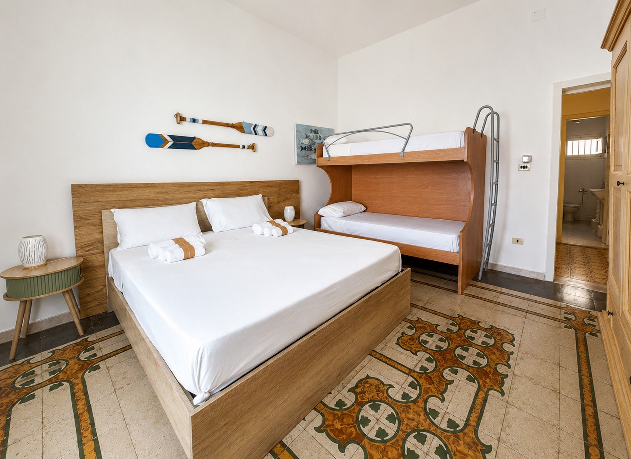 A hotel room with a large bed, a smaller bed, a bunk bed, a wooden headboard, a patterned rug, and a painting on the wall.