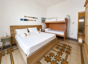 Une chambre d’hôtel avec un grand lit, un lit plus petit, un lit superposé, une tête de lit en bois, un tapis à motifs et un tableau au mur.