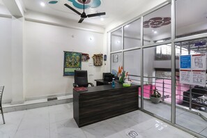 A reception desk with a dark wood finish, a ceiling fan, and a glass partition with informational posters. - Hotel O by OYO Rajdhani Hotel (Lucknow)