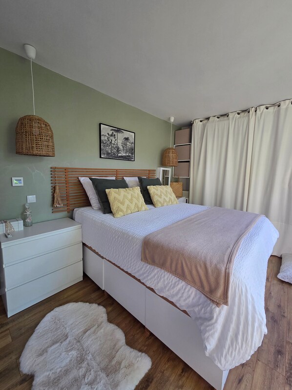 Quarto com cama, mesas de cabeceira, luminária pendente, prateleira na parede e janela com cortinas.