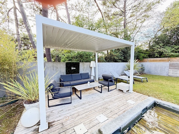 Un espace extérieur aménagé, abrité et doté d’un canapé, de chaises et d’une table, entouré de verdure et bordé d’un mur en béton.
