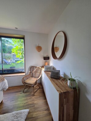 Une pièce avec une grande fenêtre, un fauteuil en osier, un banc en bois, un miroir et une plante en pot.