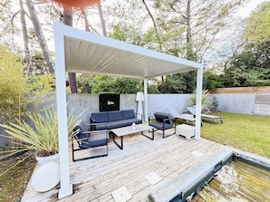 A covered outdoor seating area with a sofa, chairs, and a table, surrounded by greenery and a concrete wall. - Bed and Breakfast 'Maison Océâme - Chambre 2' with Shared Pool, Wi-Fi and Air Conditioning (Capbreton)
