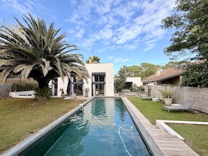 Uma área de piscina moderna, com céu límpido, uma palmeira e uma casa com grandes janelas.