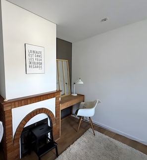 Una sala de estar con chimenea, una silla blanca, un escritorio con lámpara y una cita enmarcada en la pared.