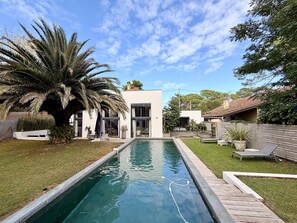 Uma área de piscina moderna, com céu límpido, uma palmeira e uma casa com grandes janelas.