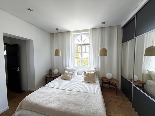Quarto com uma cama grande, duas mesas de cabeceira, um espelho e uma janela com cortinas.