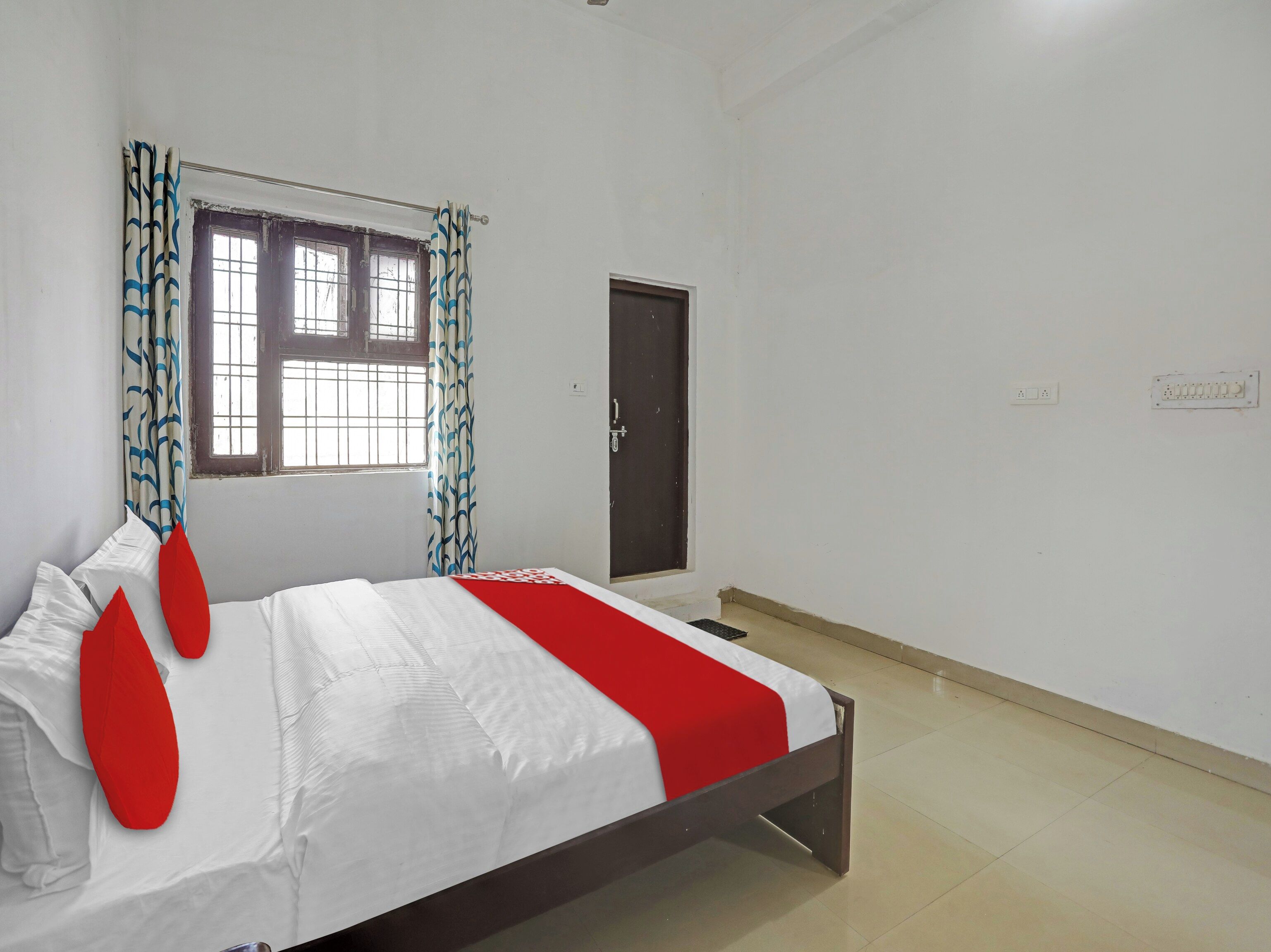 Un dormitorio con cama, ventana con cortinas, una puerta y un interruptor de luz en la pared.