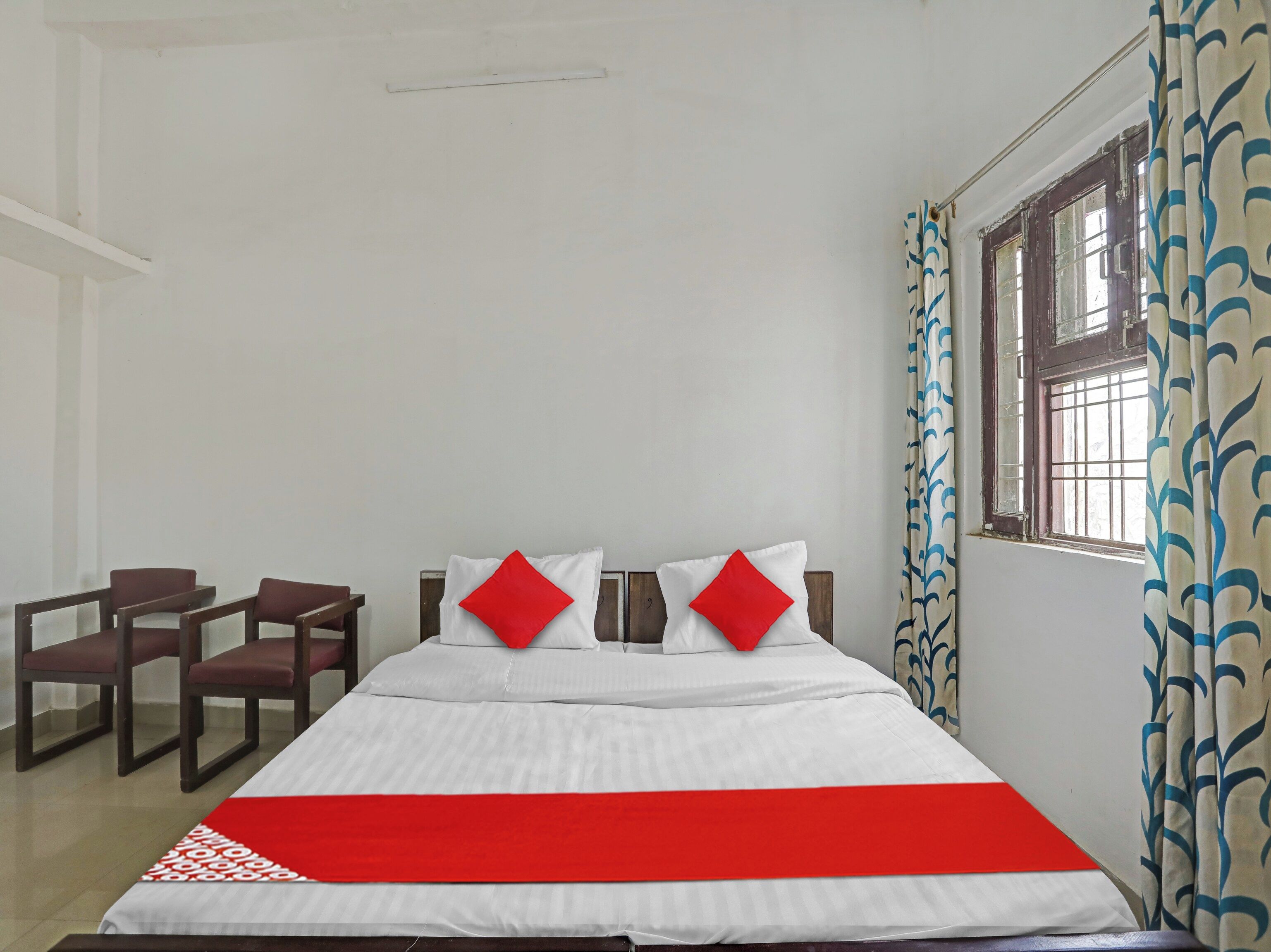 Una habitación de hotel con cama, dos sillas, una ventana con cortinas y una colcha roja.