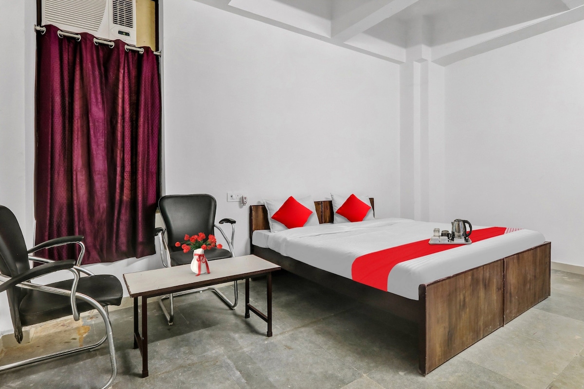 A single bed with a wooden frame, a small table with a vase of flowers, a chair, and a red and white bedspread.