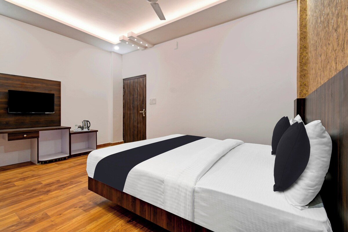 A modern hotel room with a wooden floor, a bed with white and black bedding, a flat-screen TV mounted on the wall, and a small desk with a kettle.