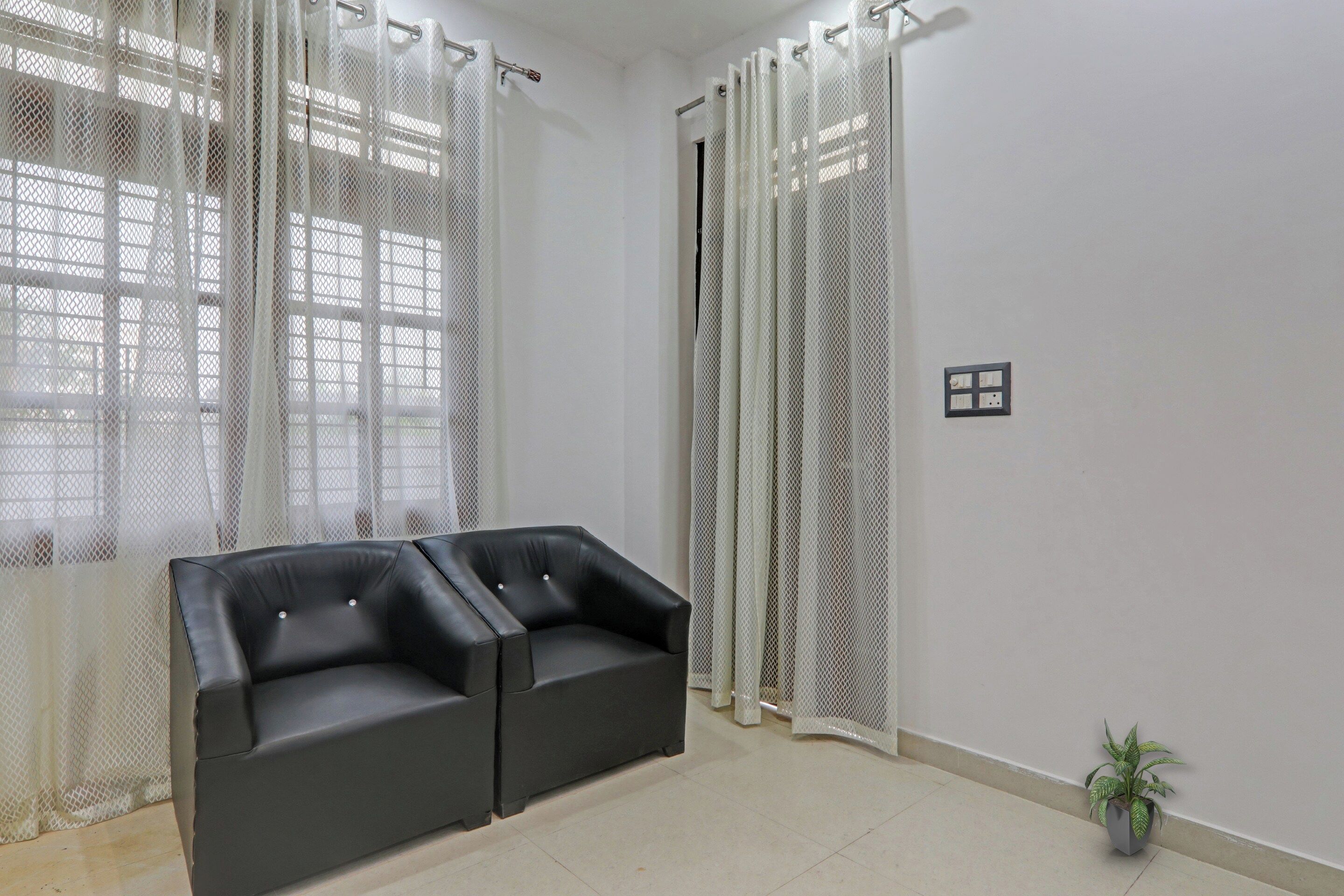 Una habitación con un sofá de cuero negro, cortinas blancas, una planta en maceta y un enchufe en la pared.
