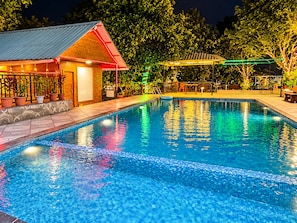 Una piscina con borde de azulejos, rodeada de plantas en macetas y un edificio con tejado rojo.