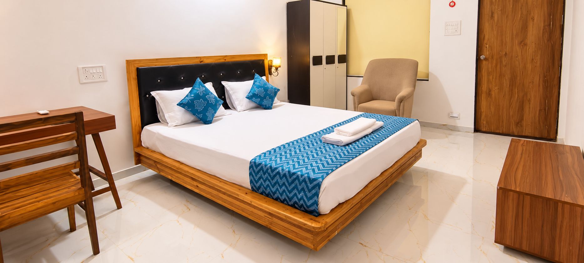 Una cama bien hecha con estructura de madera, dos almohadas y una colcha azul estampada.