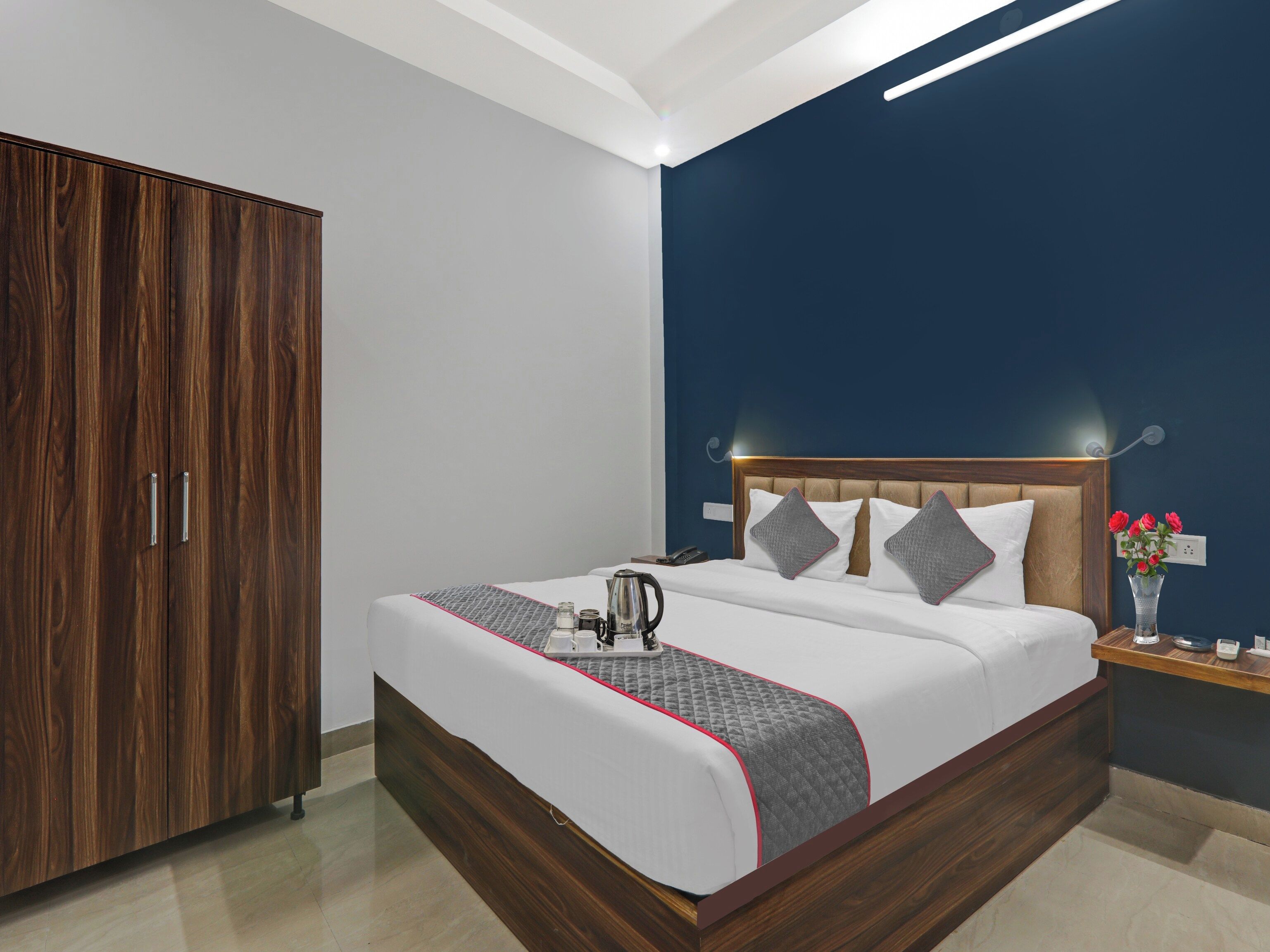 A hotel room with a wooden wardrobe, a bed with a quilted blanket, a bedside table with a vase of flowers, and a nightstand with a telephone.