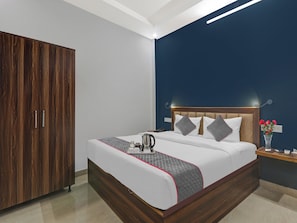 A hotel room with a wooden wardrobe, a bed with a quilted blanket, a bedside table with a vase of flowers, and a nightstand with a telephone. - Townhouse swaroop inn (Lucknow)