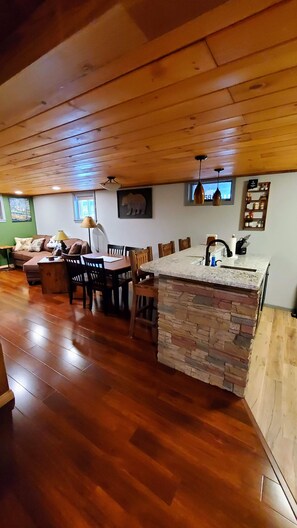 Ett mysigt vardagsrum med ett träpanelat tak, ett matbord med stolar, en köksö med en väggbeklädnad av sten och en grön vägg med en tavla.