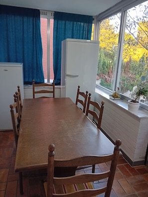 Een eetgedeelte met een houten tafel en stoelen, een witte koelkast, en een raam met gordijnen.