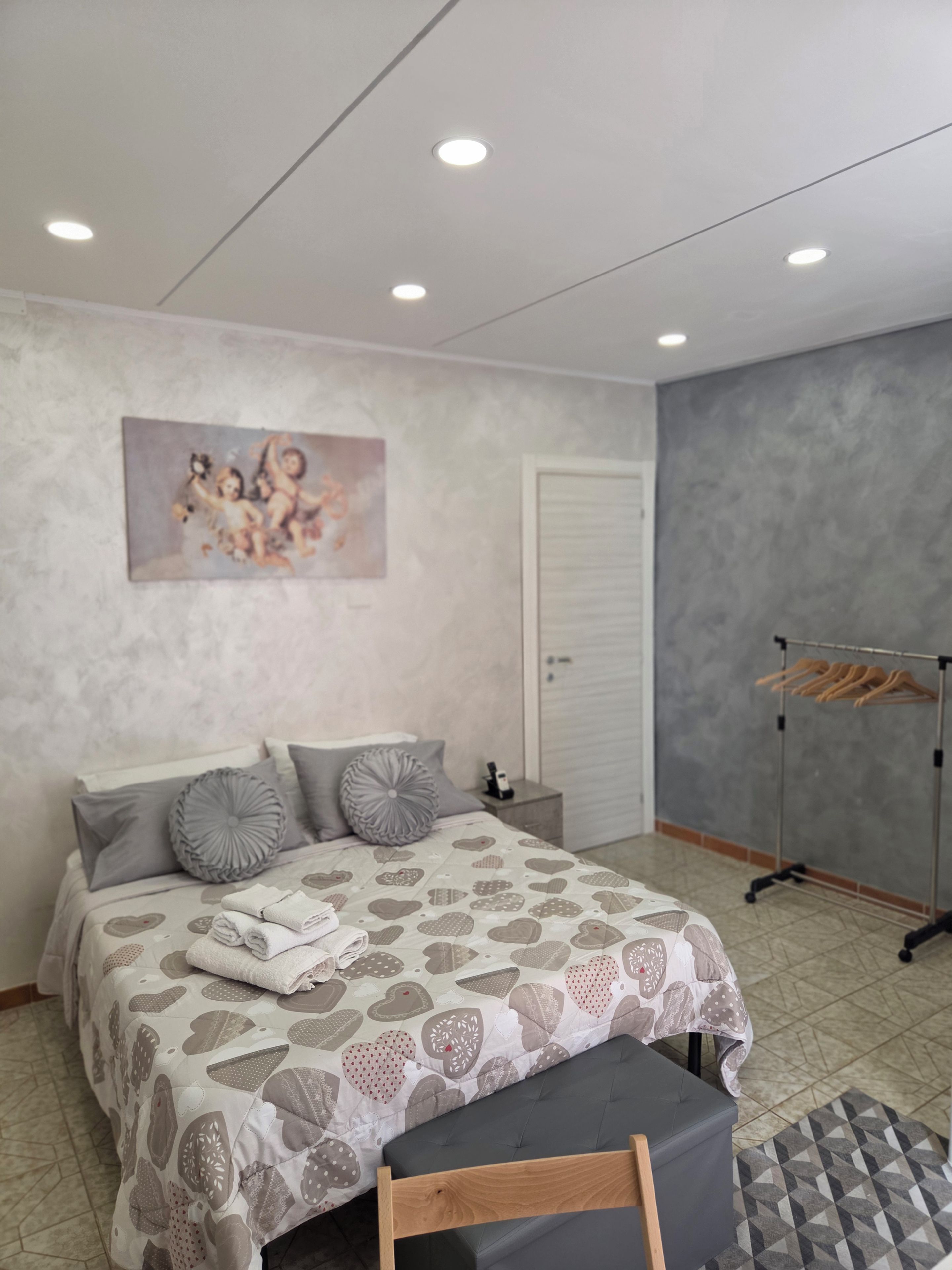 Un dormitorio con cama, almohadas, una mesita de noche, un perchero y un cuadro en la pared.