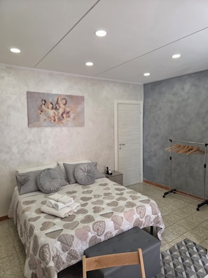 Un dormitorio con cama, almohadas, una mesita de noche, un perchero y un cuadro en la pared.