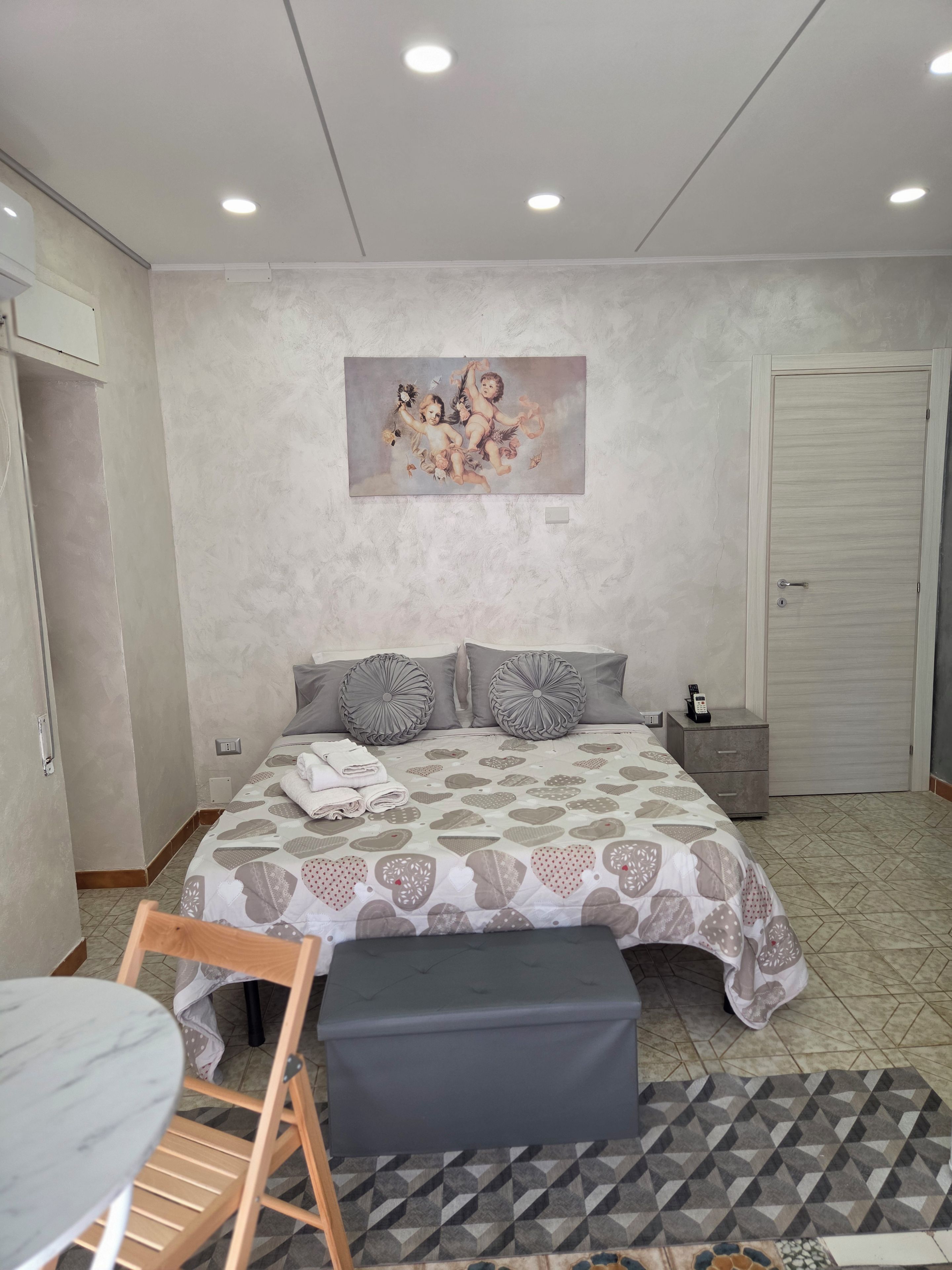 Un dormitorio con cama, mesitas de noche, un cuadro, una silla y una pequeña mesa.