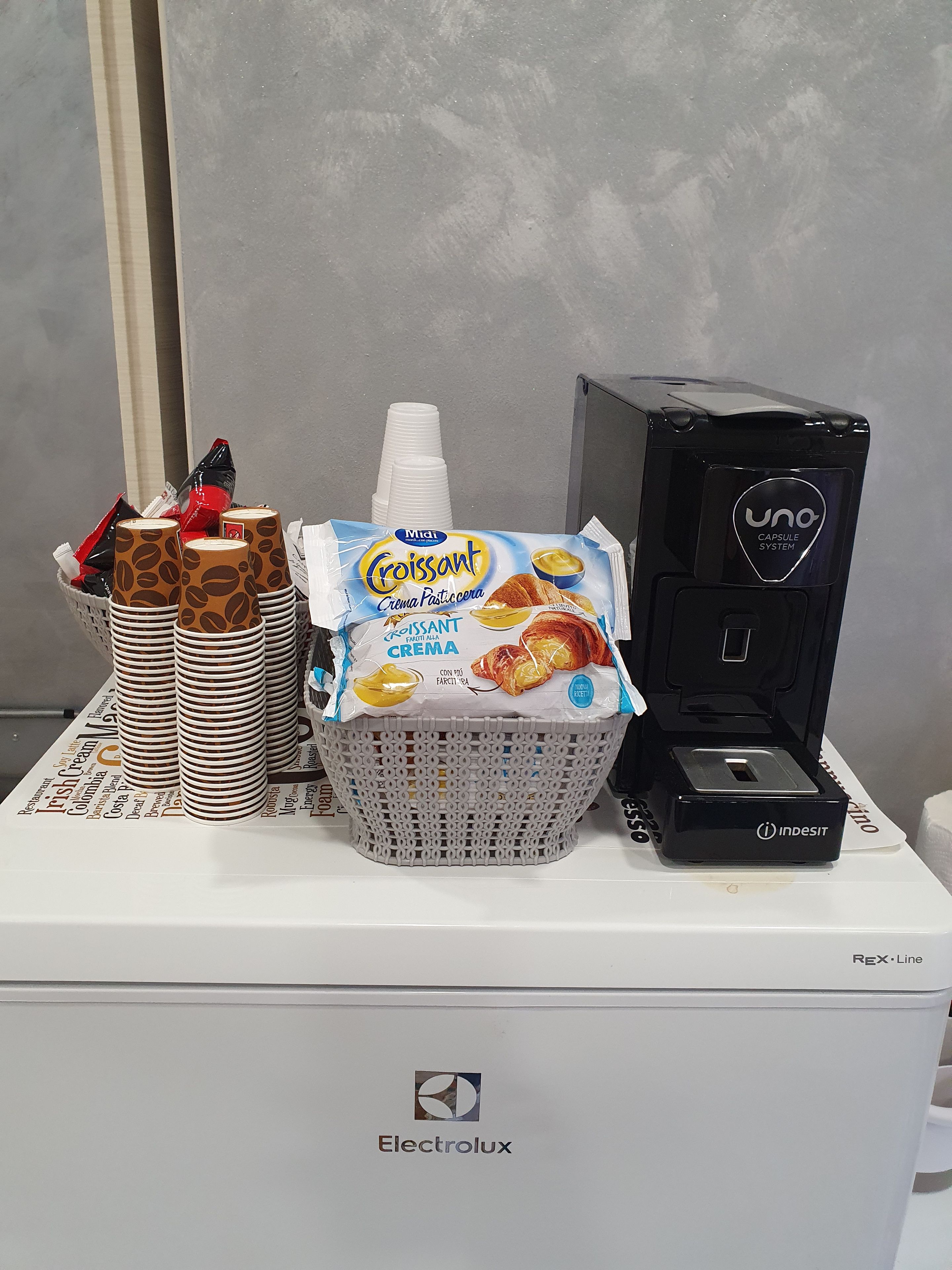 Eine Kaffeemaschine, eine Korb mit Croissantverpackungen und ein Stapel Papierbecher auf einer weißen Fläche.