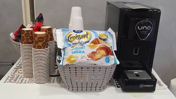 Eine Kaffeemaschine, eine Korb mit Croissantverpackungen und ein Stapel Papierbecher auf einer weißen Fläche.