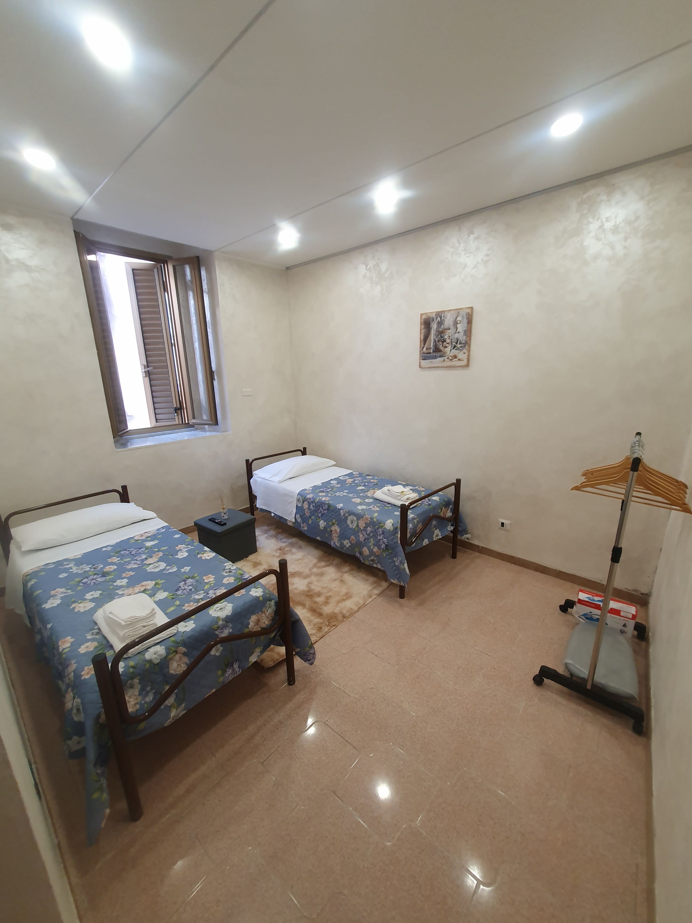 Ein Zimmer mit zwei Einzelbetten, einem Fenster, einem Bild an der Wand und einem Standventilator.