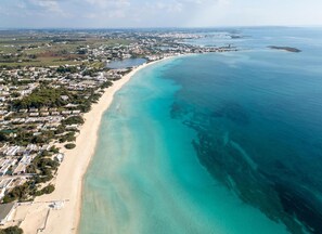 Ein Luftbild einer Küstenregion mit einem langen Sandstrand, klarem türkisblauem Wasser und einer dicht bebauten Wohngegend mit Häusern und Gebäuden.