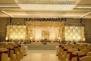 A formal event space with rows of chairs, a stage with floral decorations, and a chandelier. - POP 84974F Hotel Tulips Grand (Hyderabad)