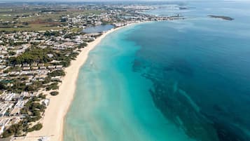 Ein Luftbild einer Küstenregion mit einem langen Sandstrand, klarem türkisblauem Wasser und einer dicht bebauten Wohngegend mit Häusern und Gebäuden.